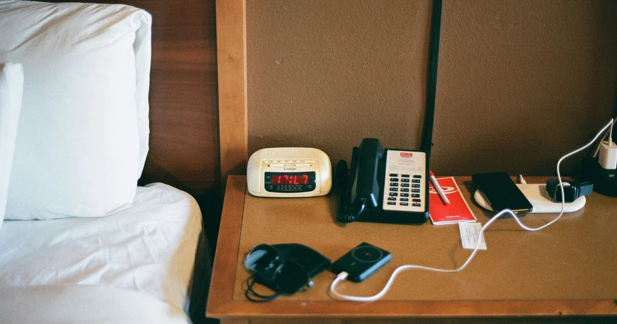 Mesa de cabeceira de hotel com telefone, relógio, aparelhos e cabos ao lado da cama.